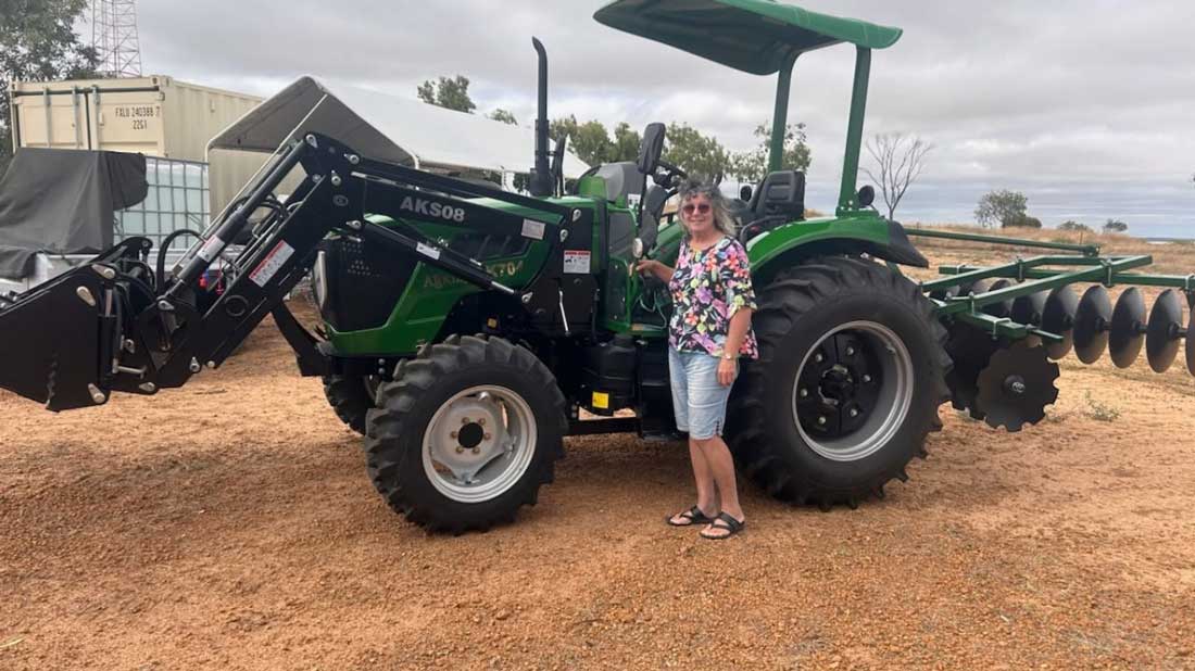 Common implement names AgKing AK508 tractor with a disc harrow attached, woman standing beside it in an outdoor farm/sale yard setting.